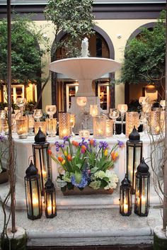 a table with candles and flowers on it in front of a fountain surrounded by potted plants