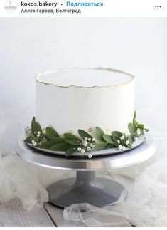 a white cake with greenery on top is sitting on a silver platter in front of a white wall