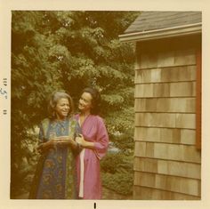 two women standing next to each other in front of a house with trees and bushes