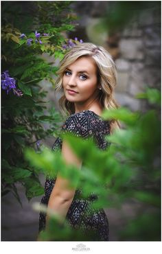 a beautiful blonde woman standing in front of purple flowers and greenery looking at the camera