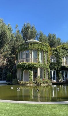 a woman is standing in front of a house with birds flying around the pond and trees behind her