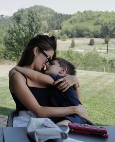 a woman holding a small child while sitting on top of a bench in the grass