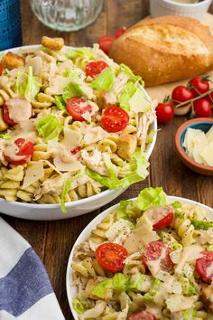 two bowls filled with pasta salad on top of a table next to bread and tomatoes