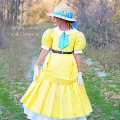 a woman in a yellow dress and hat walking down a dirt road with trees behind her