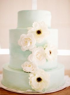 a three tiered cake with white flowers on the top and green icing, sitting on a wooden table