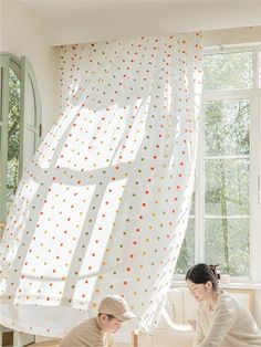 two people sitting on the floor in front of a window with polka dot curtains behind them