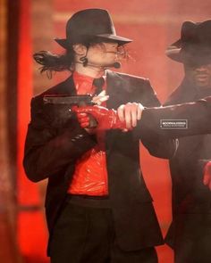 two men in black suits and red shirts are performing on stage with one man wearing a top hat