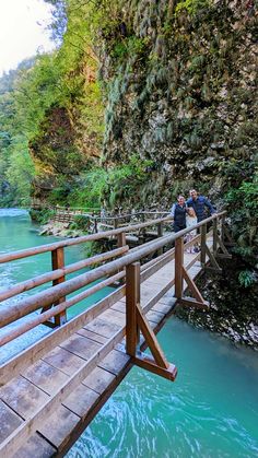 two people standing on a wooden bridge over a body of blue water in the mountains