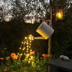 a watering can is lit up with fairy lights in the middle of a flower garden