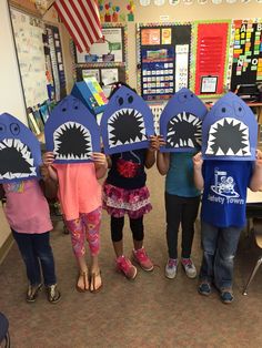 four children holding up paper shark masks in a classroom