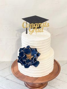 a white cake topped with a blue flower and a gold graduation hat topper that reads congrats grad