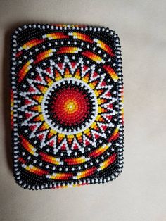 a beaded square with an orange, yellow and red design on the center is sitting on a white surface