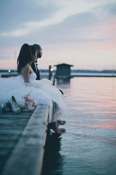 a woman in a wedding dress sitting on a dock next to the water at sunset