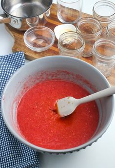 a white bowl filled with red liquid next to other glasses and spoons on a wooden cutting board