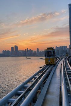 a yellow train traveling over a bridge next to the ocean at sunset with city buildings in the background