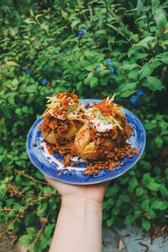 a hand holding a blue plate with food on it in front of some bushes and flowers