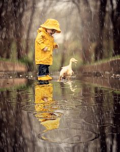 a little boy in yellow raincoat and hat standing in water with his duckling