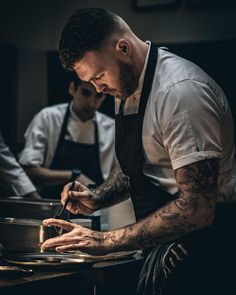 two men in aprons are preparing food at a restaurant kitchen counter with one man holding a spoon and the other is looking down