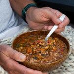 a person holding a bowl of soup with a spoon in it while sitting on a blanket