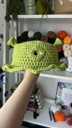 a hand holding up a green crocheted hat in front of shelves filled with yarn