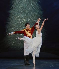 a man and woman dressed in traditional ballet attire, dancing with a christmas tree behind them