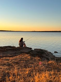 a woman sitting on top of a rock next to the ocean with a fishing rod