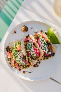 three tacos are sitting on a white plate with a lime and some other food