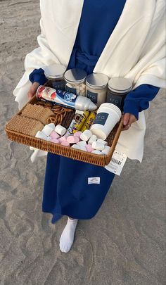 a woman holding a wicker basket filled with various items on the beach in front of her