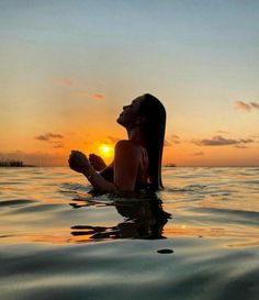 a woman sitting in the water at sunset with her hands together and looking up into the sky