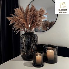 three candles sit on a table in front of a round mirror and some dried plants