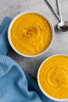 two white bowls filled with yellow soup on top of a blue towel next to spoons