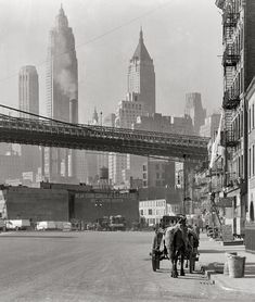 an old black and white photo of people riding horses down the street with tall buildings in the background