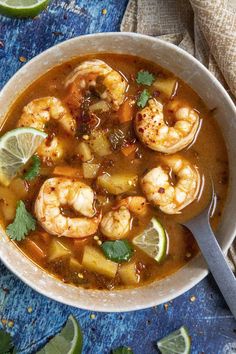 a bowl of soup with shrimp, lime and cilantro on the side next to a spoon