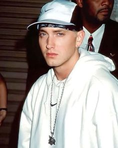 a young man wearing a white hat and chain around his neck with another man in the background