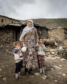 a woman standing next to two children in front of a stone building with sheep behind her