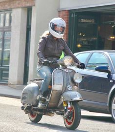 a woman riding on the back of a silver scooter down a city street