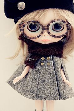 a doll with big eyes wearing a black hat and gray dress is standing in front of a white wall