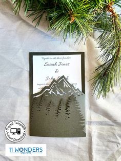 a wedding card with mountains and pine trees on it, sitting next to a christmas tree