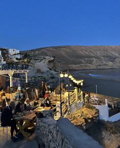 people sitting at tables on the side of a cliff overlooking a body of water with buildings in the background