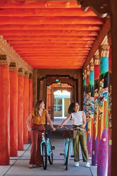 two women riding bikes in an orange covered walkway with colorful columns on either side and one woman standing next to the bike