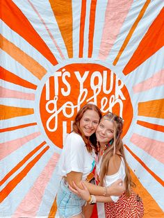 two young women hugging each other in front of a large orange and white banner that says it's your joy