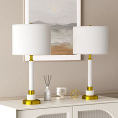 two lamps are sitting on top of a white cabinet in front of a wall with a painting