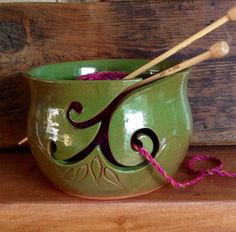 a green bowl with two knitting needles in it sitting on a wooden shelf next to yarn