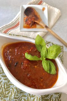 a white bowl filled with tomato sauce and basil on top of a green napkin next to a wooden spoon