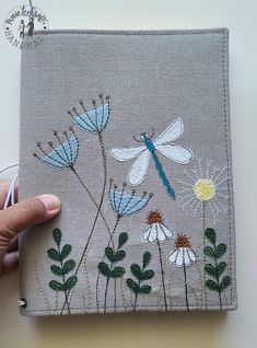 a hand is holding an embroidered book with dragonflies and daisies on it