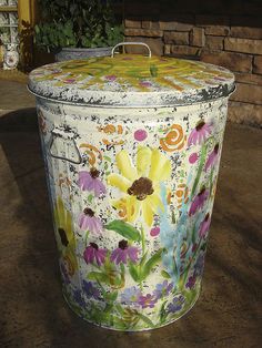 a painted trash can sitting on the ground next to a brick wall and potted plant