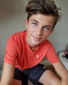 a young boy sitting on top of a bed wearing a red shirt and black shorts