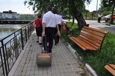 two people walking with luggage down a sidewalk next to a bench and water in the background