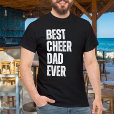 "Show your support for your cheerleader with this \"Cheer Dad\" shirt. Perfect gift for proud fathers of cheerleaders. Comfortable and stylish tee that's great for games, events, and everyday wear. The unisex heavy cotton tee is the basic staple of any wardrobe. It is the foundation upon which casual fashion grows. All it needs is a personalized design to elevate things to profitability. The specially spun fibers provide a smooth surface for premium printing vividity and sharpness. No side seams mean there are no itchy interruptions under the arms. The shoulders have tape for improved durability. .: 100% cotton (fiber content may vary for different colors) .: Medium fabric (5.3 oz/yd² (180 g/m .: Classic fit .: Tear-away label .: Runs true to size" Father's Day Team Spirit T-shirt With Crew Neck, Father's Day Team Spirit Crew Neck T-shirt, Black Sports Fan T-shirt For Cheerleading, Father's Day Sports Fan T-shirt With Letter Print, Father's Day Cotton T-shirt With Team Spirit, Father's Day School Spirit Cotton T-shirt, Black Crew Neck T-shirt For Cheerleading, Crew Neck T-shirt With Text Print For Cheerleading, Short Sleeve Letter Print Shirt For Cheerleading