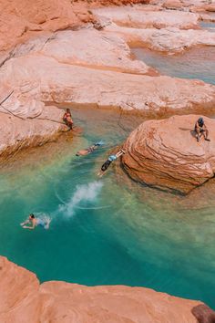 people are swimming in the water near some large rocks and sand cliffs, with one person falling off his surfboard